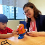 A three-year-old boy named Jaiden plays as his speech-language therapist smiles.