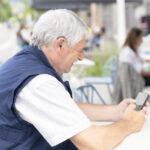 An older man with grey hair smiles as he uses his mobile phone to adjust his cochlear implant settings.