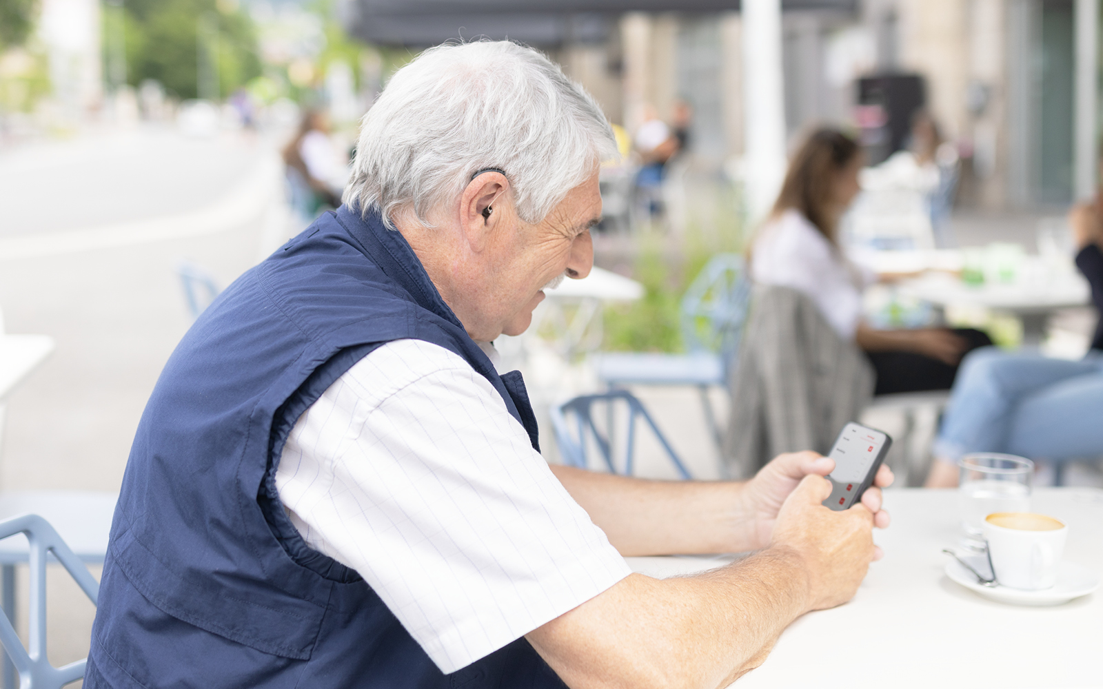 An older man with grey hair smiles as he uses his mobile phone to adjust his cochlear implant settings.