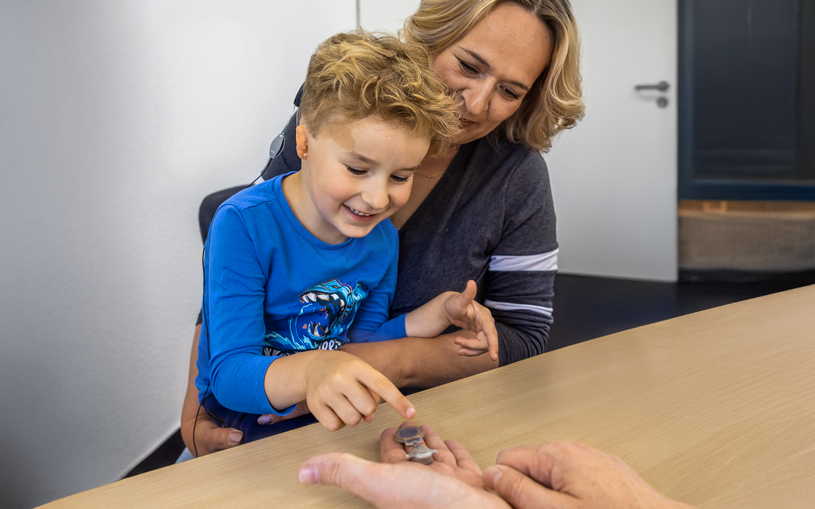 A hand holding the BONEBRIDGE BCI 602 implant is extended over a desk at a clinic as a smiling young boy with microtia reaches out to touch it as his mother holds him in her lap and looks on.