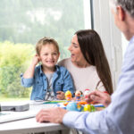 A young child sits beside an adult while a clinician adjusts and activates the child’s cochlear implant audio processor. The clinician works at a nearby desk with a laptop displaying audio settings, and colorful toys are placed on the table to engage the child. A large window behind them shows a green outdoor landscape.