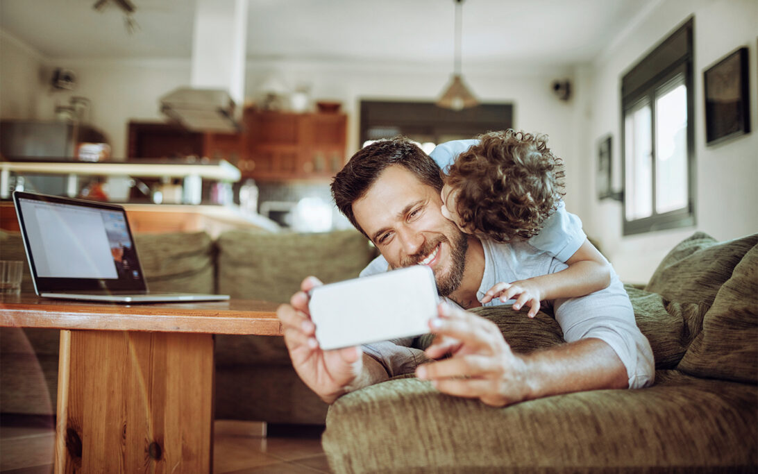 A person reclining on a couch in a living room holds up a smartphone with the littlears auditory questionnaire on it, while a small child climbs onto the person's back. A laptop sits open on a wooden coffee table nearby, and the background shows a kitchen area with warm lighting.