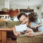 A person reclining on a couch in a living room holds up a smartphone with the littlears auditory questionnaire on it, while a small child climbs onto the person's back. A laptop sits open on a wooden coffee table nearby, and the background shows a kitchen area with warm lighting.