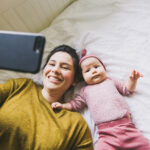 An adult and an infant are lying side‑by‑side on a bed with light-colored bedding. The adult is holding a smartphone above them, and the screen is being used to fill in the LittleEARS auditory questionnaire within the ReDi app. The infant is dressed in pink clothing and lying on their back, while the adult wears a long‑sleeved top. The background shows a simple, bright bedroom setting.