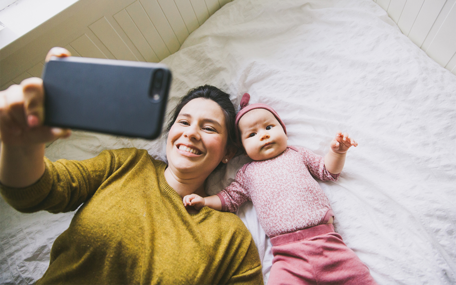 An adult and an infant are lying side‑by‑side on a bed with light-colored bedding. The adult is holding a smartphone above them, and the screen is being used to fill in the LittleEARS auditory questionnaire within the ReDi app. The infant is dressed in pink clothing and lying on their back, while the adult wears a long‑sleeved top. The background shows a simple, bright bedroom setting.