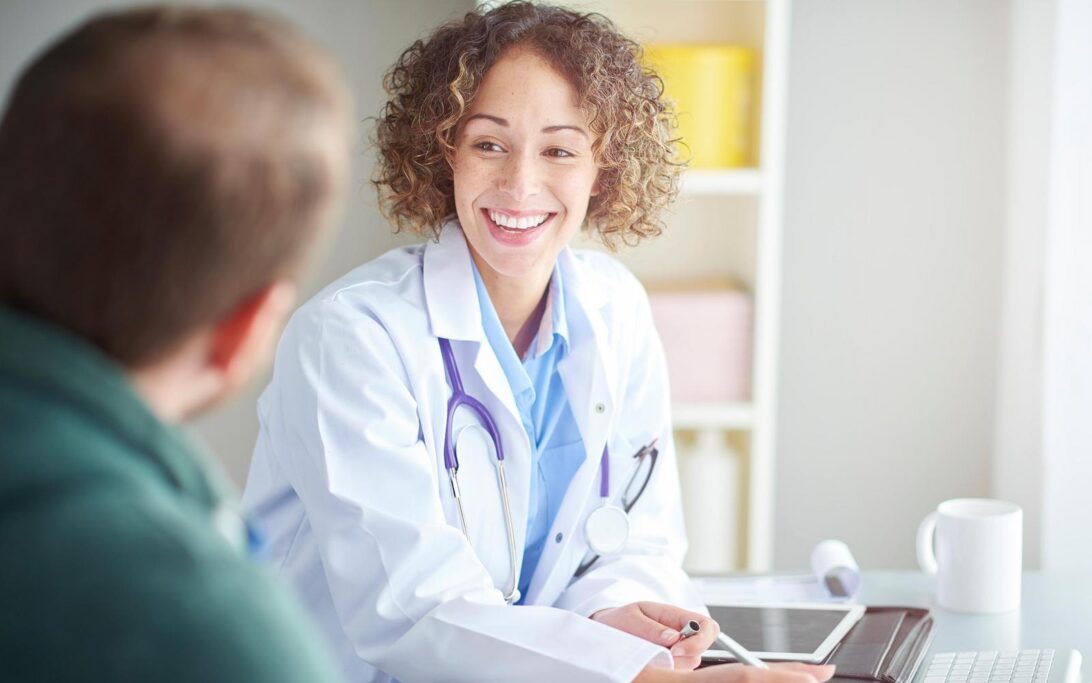 A medical professional wearing a white lab coat and stethoscope sits at a desk, speaking with a hearing loss patient who is seated across from them. The doctor holds a pen over a clipboard with papers, which is a quality of life questionnaire. A tablet and an open laptop rest on the desk. A white cup and various office items are visible in the background, creating the setting of a bright, modern medical consultation room.
