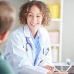 A medical professional wearing a white lab coat and stethoscope sits at a desk, speaking with a hearing loss patient who is seated across from them. The doctor holds a pen over a clipboard with papers, which is a quality of life questionnaire. A tablet and an open laptop rest on the desk. A white cup and various office items are visible in the background, creating the setting of a bright, modern medical consultation room.