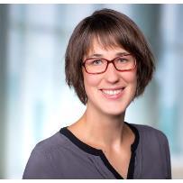 A woman with short, straight brown hair, wearing a dark grey top with a subtle seam detail near the neckline, standing in front of a softly blurred background of cool blue and white tones in an indoor, well‑lit professional setting.
