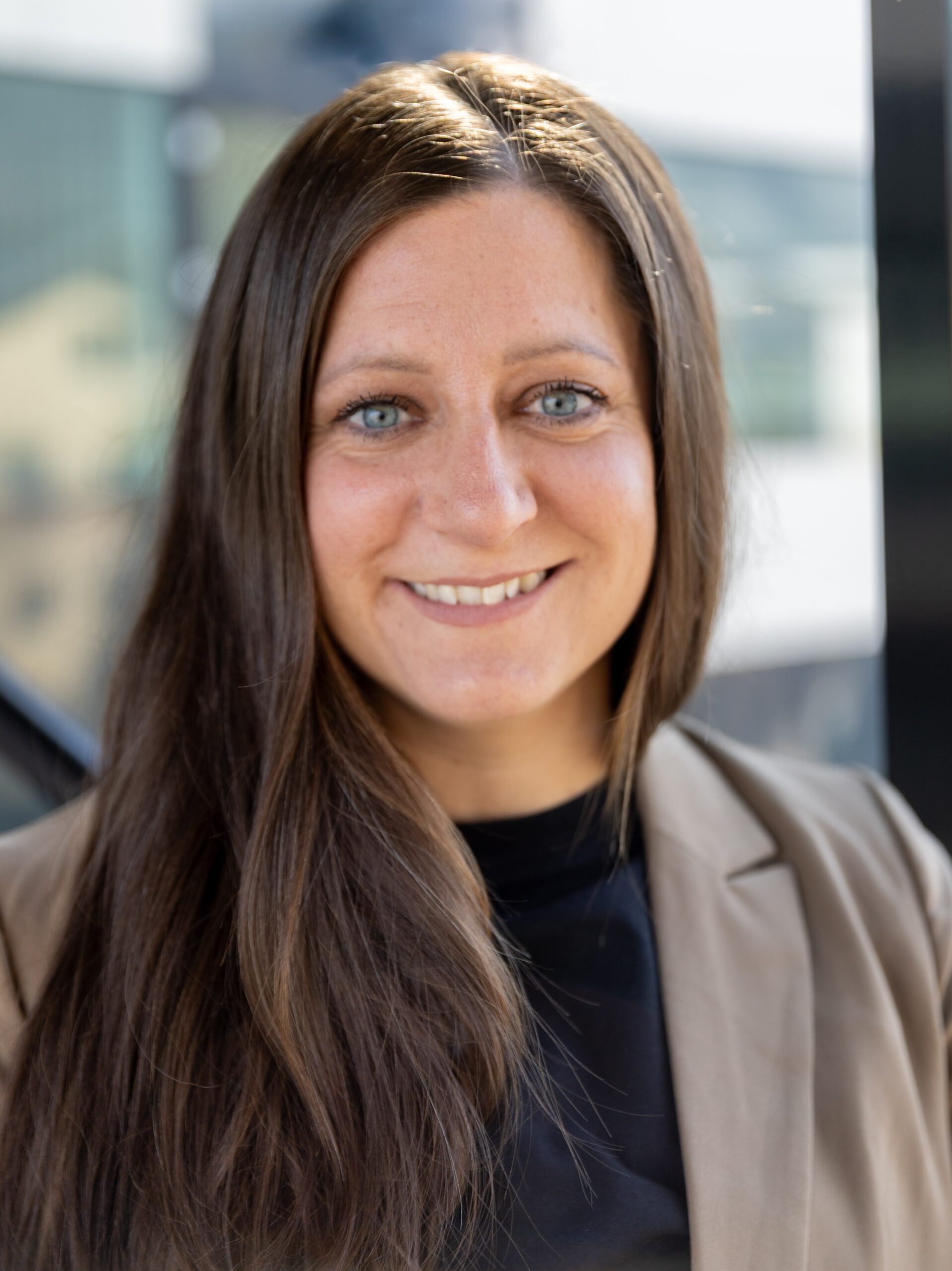 Head-and-shoulders portrait of Sandra Eisner, a Speech-Language Pathologist from Austria, looking at the camera. Sandra Eisner has long brown hair worn loose and is dressed in a dark top with a light-colored blazer. The background is softly blurred, suggesting an indoor professional setting.