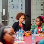 Group of people seated around a table during a meeting or workshop, with several water bottles, notebooks, and smartphones visible. Two participants in the center look down at their devices and papers, while name cards—one reading “Adelaide” and another “berhane”—are placed on the table. The setting appears to be a conference room with glass doors featuring a geometric patterned design and a visible “PULL” sign on the door.