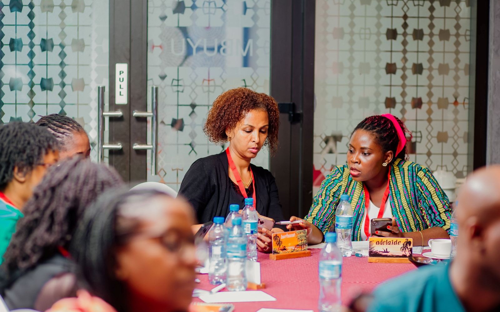 Group of people seated around a table during a meeting or workshop, with several water bottles, notebooks, and smartphones visible. Two participants in the center look down at their devices and papers, while name cards—one reading “Adelaide” and another “berhane”—are placed on the table. The setting appears to be a conference room with glass doors featuring a geometric patterned design and a visible “PULL” sign on the door.