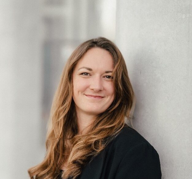 Portrait of a person with long hair wearing a black blazer, standing beside a concrete wall in a bright hallway.