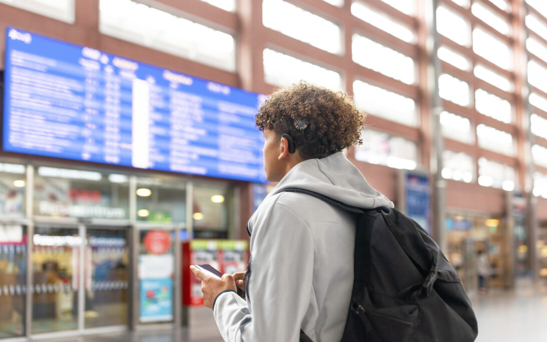 Teenager with a cochlear implant checks a departure board in a transit terminal using a smartphone.