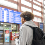 Teenager with a cochlear implant checks a departure board in a transit terminal using a smartphone.